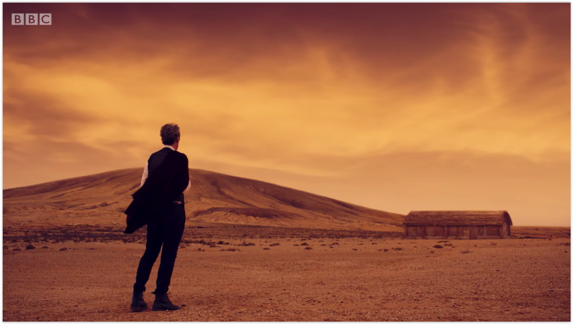 A screencap from 'Hell Bent'. A wide shot of the Doctor looking over at the barn on Gallifrey against an amber-orange sky.