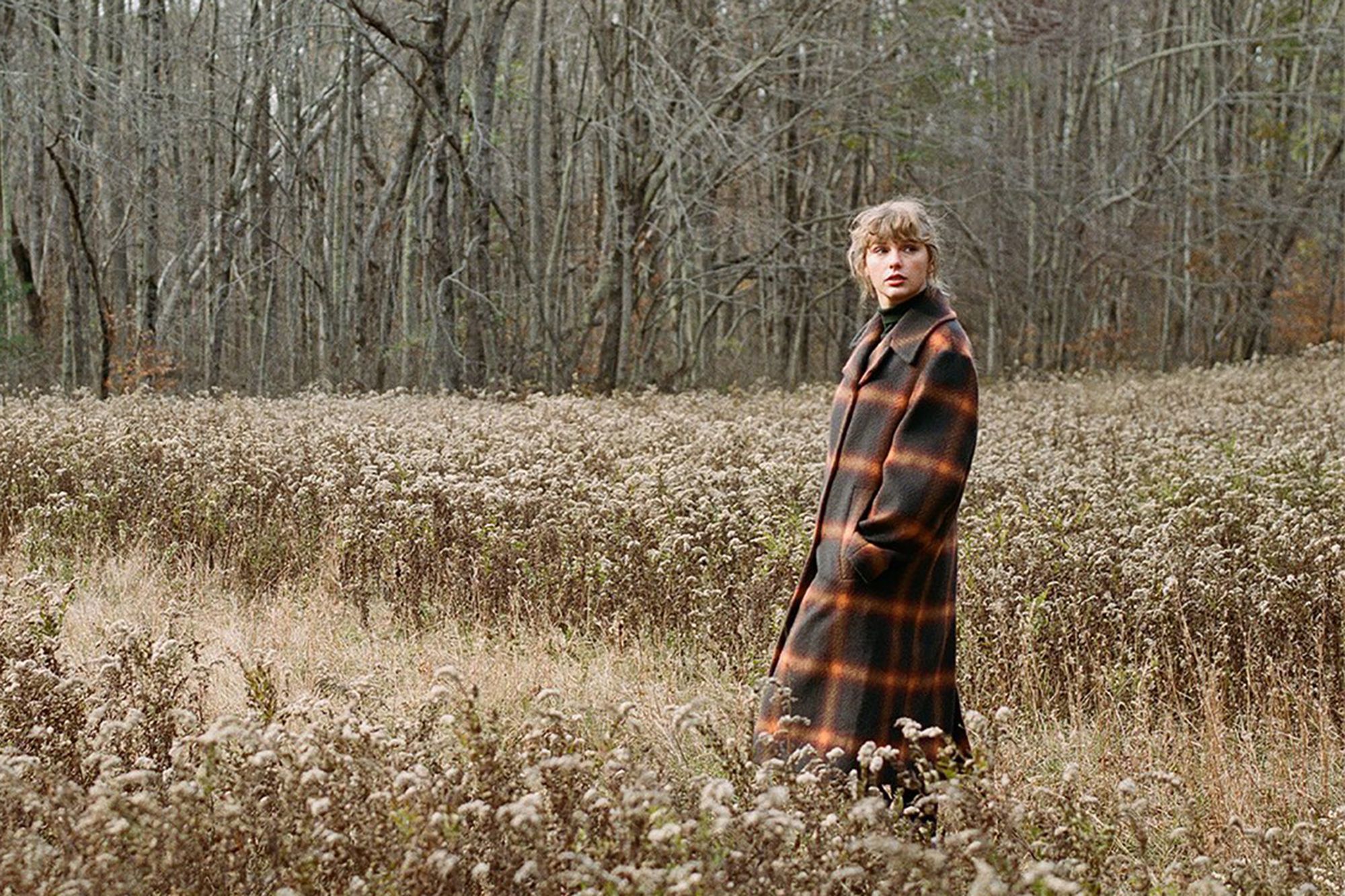 A promotional shot for the Taylor Swift album Evermore. Taylor Swift looking pensive wearing a flannel coat in an overgrown field with barren trees in the background, from the promotional shots for Evermore.